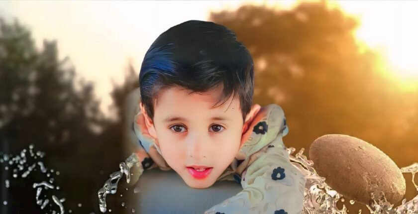 A young child with short, dark hair is lying on the ground in a natural outdoor setting. The background features trees and a warm, golden light, suggesting it might be sunrise or sunset. The child is wearing a light-colored jacket with floral patterns and has one hand resting near their face. The overall scene conveys a peaceful and serene atmosphere