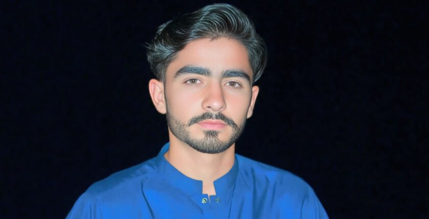 A young man with neatly styled dark hair and a trimmed beard is shown in the image. He is wearing a bright blue shirt with a traditional collar, featuring small decorative buttons. The background is dark, which contrasts with his attire and highlights his facial features. His expression is neutral, and he is looking directly at the camera